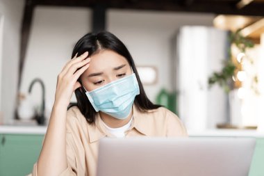Tıbbi maske takmış yorgun Asyalı kız Webinar dinliyor, laptopta sorunlu yüzüyle smth izliyor, ödev yapıyor..