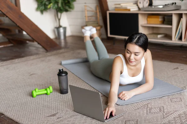 Asian fitness girl taking online fitness course, watching sports video, exercising at home on floor mat with dumbells and protein shaker bottle, smiling at camera.