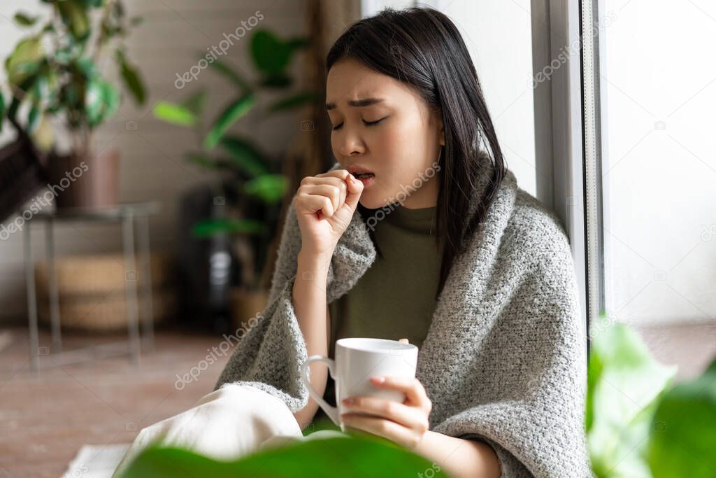 Sad ill asian girl staying on self quarantine during covid, catching flu and sitting at home with cup of tea near window, coughing in hand.