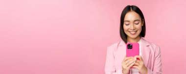 Gülümseyen iş kadını portresi, akıllı telefon kullanan Asyalı şirket çalışanı, cep telefonu uygulaması, pembe arka planda duran.