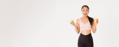 Spor, refah ve aktif yaşam tarzı konsepti. Heyecanlı Asyalı fitness kızı, elma ve portakal suyu içen sporcu kadın şaşırmış ve mutlu, sağlıklı beslenmek için zinde kalmak için, beyaz arka plan..
