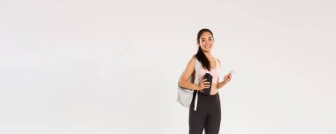 Sağlıklı gülümseyen, zayıf Asyalı kız fitness antrenmanına gidiyor, bayan sporcu spor aletleri ve su şişesiyle sırt çantası taşıyor, cep telefonu spor uygulaması kullanıyor, beyaz arka plan kullanıyor..