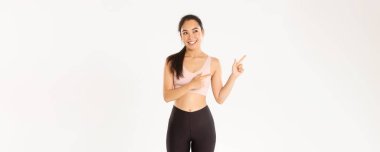 Spor, refah ve aktif yaşam tarzı konsepti. İlgi çekici bir gülümseme, güzel bir Asyalı fitness kızı, sporcu bir kadın sol üst köşeyi işaret ediyor ve arıyor, düşünceli, seçim yapıyor, spor ekipmanı seçiyor..