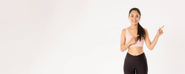 Spor, refah ve aktif yaşam tarzı konsepti. Gülümseyen ve zayıf kadın fitness eğitmeni, spor salonunda çalışan Asyalı kız ve bir şey gösteren, parmaklarını sağa doğru gösteren, beyaz arka planda duran.