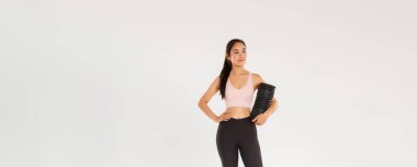 Gülümseyen, kendinden emin ve motive olmuş Asyalı bayan jimnastik koçu, spor giyim fitness kızı, etrafa bakan, idmandan sonra kullanmak için köpük silindir tutan, eğitim ekipmanlarıyla ayakta duran..