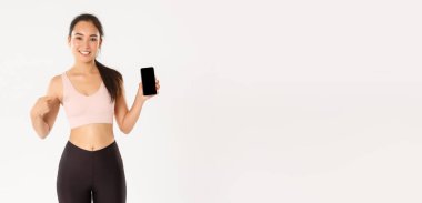 Spor, teknoloji ve aktif yaşam tarzı konsepti. Tatmin olmuş gülümseyen Asyalı fitness kızı, çekici sporcu kadın akıllı telefon ekranını işaret eden, mobil egzersiz takip uygulaması öneren.