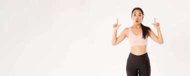 Spor, refah ve aktif yaşam tarzı konsepti. Heyecanlı ve mutlu Asyalı spor giyim kızı, parmaklarını havaya kaldırıyor ve büyük indirimlerle şaşkın bakıyor, afiş okuyor, beyaz arka plan.