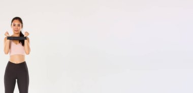 Gülümseyen ve kararlı, zayıf esmer Asyalı fitness kızı, antrenman koçu dirençli halatla egzersiz yapıyor, kameraya bakıyor, egzersiz ekipmanlarını gösteriyor..