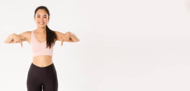Spor, refah ve aktif yaşam tarzı konsepti. Gülümseyen ve zayıf kadın fitness eğitmeni, spor salonunda çalışan Asyalı kız ve bir şey gösteren, parmaklarını yere doğru gösteren, beyaz arka planda duran.