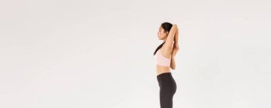 Odaklanmış güçlü Asyalı kadın koçun tam boy profili, spor yapan sporcu kadın spor yapıyor, ellerini esnetiyor, kollarını arkada kenetliyor, beyaz arka planda duruyor..