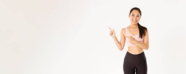 Spor, refah ve aktif yaşam tarzı konsepti. İlgi çekici bir gülümseme, güzel bir Asyalı fitness kızı, sporcu bir kadın sol üst köşeyi işaret ediyor ve arıyor, düşünceli, seçim yapıyor, spor ekipmanı seçiyor..