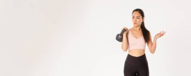 Spor, refah ve aktif yaşam tarzı konsepti. Aktif kıyafetli kafası karışmış Asyalı fitness kızı şaşkın görünüyor, kettlebell kaldırıyor, egzersiz ve sağlıklı beslenme ile güçleniyor, beyaz arka planda duruyor..
