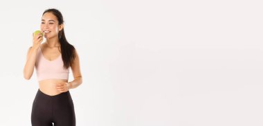 Spor, refah ve aktif yaşam tarzı konsepti. Tatmin olmuş gülümseyen Asyalı fitness kızı yeşil elmayı zevkle yiyor, spor salonundaki verimli egzersizden sonra memnun görünüyor, beyaz arka plan..