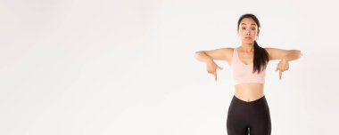 Spor, refah ve aktif yaşam tarzı konsepti. Şaşırmış ve etkilenmiş Asyalı fitness kızı, spor salonu üyesi ya da aktif giyimli kadın parmakları aşağıda, nutku tutulmuş, beyaz arkaplan.