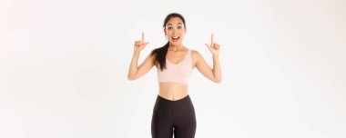 Spor, refah ve aktif yaşam tarzı konsepti. Heyecanlı ve mutlu Asyalı spor giyim kızı, parmaklarını havaya kaldırıyor ve büyük indirimlerle şaşkın bakıyor, afiş okuyor, beyaz arka plan.