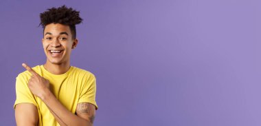 Promosyon gösteren neşeli, çekici genç erkek öğrencinin yakın plan portresi, sol üst köşeye işaret ediyor, gülümsüyor memnun, tıklama linki tavsiye ediyor, ziyaret sayfası veya indirme uygulaması, mor arkaplan.