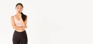 Spor, refah ve aktif yaşam tarzı konsepti. Düşünceli ince ve formda Asyalı fitness kızı spor salonunu seçiyor, düşünüyor ya da karar veriyor, kameraya bakıp eğleniyor ve aktif giysinin içinde duruyor..