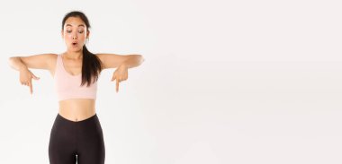 Spor, refah ve aktif yaşam tarzı konsepti. Etkilenmiş, dili tutulmuş Asyalı bir kız, fitness öğretmeni ya da jimnastik hocası, parmaklarını şaşkın bir şekilde aşağıya gösteren bir üye, iyi bir tanıtım teklifi, beyaz bir geçmiş..