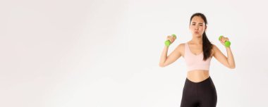 Fitness, sağlıklı yaşam tarzı ve refah konsepti. Spor kıyafetli yorgun Asyalı bir kızın portresi, antrenman sırasında bitkin görünüyor, evde online koçla egzersiz yapıyor, dambılları kaldırıyor, beyaz arka plan.