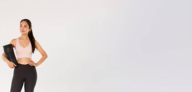 Gülümseyen, kendinden emin ve motive olmuş Asyalı bayan jimnastik koçu, spor giyim fitness kızı, etrafa bakan, idmandan sonra kullanmak için köpük silindir tutan, eğitim ekipmanlarıyla ayakta duran..