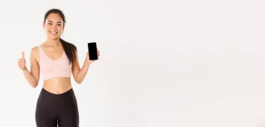 Spor, teknoloji ve aktif yaşam tarzı konsepti. Tatmin olmuş gülümseyen Asyalı fitness kızı, çekici sporcu kadın başparmak kaldıran ve akıllı telefon ekranı gösteren, mobil egzersiz takip uygulaması önerin.