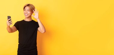 Dost canlısı yakışıklı Asyalı bir adamın portresi, gülümseyen ve akıllı telefon kamerasına el sallayan, video görüşmesinde arkadaşlarını selamlayan, sarı arka planda duran.