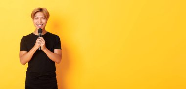 Karizmatik gülümseyen Asyalı adamın portresi mikrofon tutuyor ve karaoke söylüyor, şarkı söylüyor, sarı arka planda duruyor..