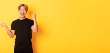 Memnun ve mutlu çekici Koreli adam gülümsüyor, sevinçle yumruk sallıyor, elinde bir şey tutuyor, sarı arka planda duruyor..