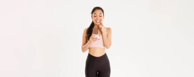 Spor, refah ve aktif yaşam tarzı konsepti. Belirsiz Asyalı fitness kızı spor giyim sektöründe çekici çikolatalara bakıyor ve diyetteyken, beyaz arka planda şeker yerken parmaklarını ısırıyor..