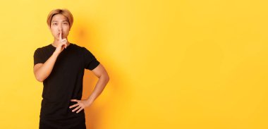 Çekici sarışın Asyalı adamın portresi şşşş, parmaklarınızı dudaklarınıza bastırın ve sessiz olun, sarı arka plan.