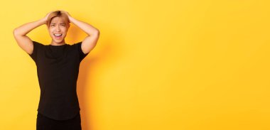 Genç Asyalı adamın portresi panikliyor, endişeli ve üzgün görünüyor, el ele tutuşuyor ve yüzünü buruşturuyor, sarı arka planda duruyor..