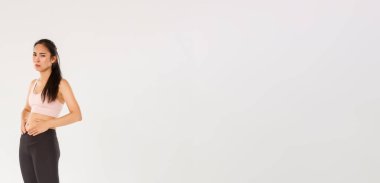 Kilo vermeye, karnına dokunmaya çalışan üzgün ve kasvetli Asyalı fitness kızı. Beyaz arka planda spor kıyafetleri içinde dururken idman ve diyet sonuçlarını göremiyor..