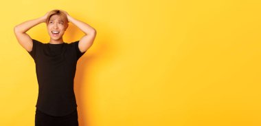 Genç Asyalı adamın portresi panikliyor, endişeli ve üzgün görünüyor, el ele tutuşuyor ve yüzünü buruşturuyor, sarı arka planda duruyor..