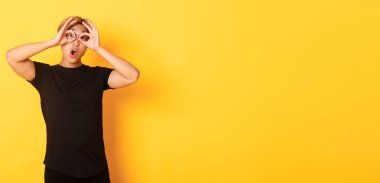 Komik ve yakışıklı Asyalı sarışının portresi parmaklarını gözlük yapıyor, yüzünü buruşturuyor, sarı arka planda duruyor..