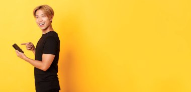 Gülümseyen yakışıklı Asyalı adamın portresi, profilde duruyor ve akıllı telefonu işaret ediyor, uygulamayı tavsiye ediyor, sarı arka planda duruyor..