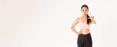 Spor, refah ve aktif yaşam tarzı konsepti. Ciddi derecede hoşnutsuz Asyalı kadın fitness eğitmeni, sporcu kadın onaylamayarak, uyararak ya da karşı çıkarak parmağını sallıyor, beyaz arka planda duruyor..