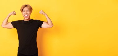 Mutlu ve kendinden emin Asyalı sarışının portresi, kaslarını esnetiyor, güçlü büyük kaslar gösteriyor, sarı arka planda duruyor..