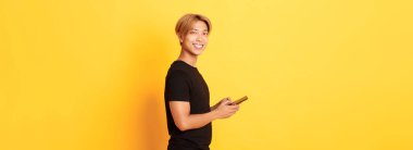 Siyah kıyafetli yakışıklı Asyalı adamın portresi, cep telefonu kullanıyor ve kameraya bakıyor, mutlu bir gülümsemeyle, sarı arka planda..