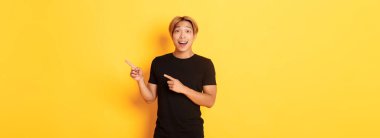 İnanılmaz yakışıklı Koreli adam, gülümsemesi büyülenmiş ve parmaklarını sağa doğru gösteriyor, yol gösteriyor, sarı arka planda duruyor..