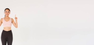 Spor, spor salonu ve sağlıklı vücut konsepti. Neşeli bir gülümseme ve hoş bir fitness kızı, parmaklarını havaya kaldırarak egzersiz malzemeleri reklamını gösteriyor, eğitim için üyelik satın alıyor..