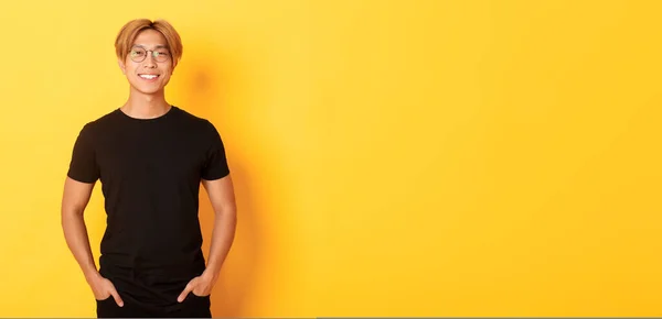 Handsome asian blond guy in glasses standing in black clothes and smiling pleased, yellow background.