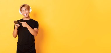 Neşeli Asyalı yakışıklı adam, müzik dinliyor ya da kulaklıkla iyi podcast yapıyor, akıllı telefonu işaret ediyor mutlu bir gülümsemeyle, sarı arka planda..