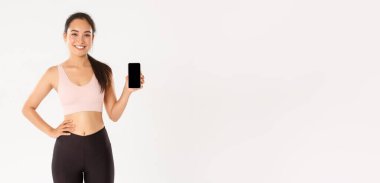 Spor, teknoloji ve aktif yaşam tarzı konsepti. Formda ve zayıf, yakışıklı Asyalı fitness kızı, spor giyim sektöründe bayan sporcu akıllı telefon ekranı ve gülümseme gösteren egzersiz uygulaması öneriyor..
