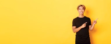 Tatmin olmuş gülümseyen sarışın adam portresi, Asyalı öğrenci parmaklarını sağa gösteriyor, yolu gösteriyor, sarı arka planda duruyor..