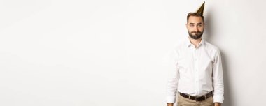 Kutlama ve tatiller. Doğum günü şapkalı kasvetli adam beyaz arka planda garip duruyor, keyifsiz hissediyor..