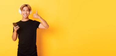 Mutlu Asyalı adam müzikten ya da podcast 'ten hoşlanır. Onaylama için onay verir, cep telefonu tutar, sarı arka planda durur..