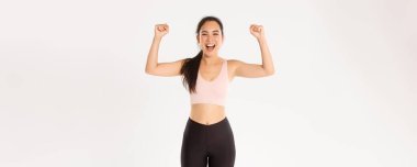 Spor, refah ve aktif yaşam tarzı konsepti. Güçlü ve güçlü Asyalı bayan sporcu, fitness kızı iyi bir egzersiz için kendini cesaretlendiriyor, yumruk tokuşturuyor ve bağırıyor, neşeli, beyaz arka plan.