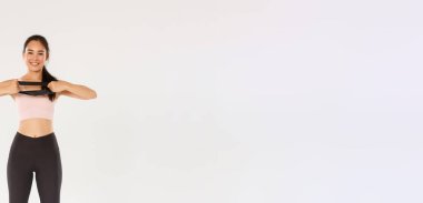 Tam boy ince ve fit görünümlü Asyalı kız spor yapıyor, direnç bandını elleriyle geriyor ve gülümsüyor. Spor salonunda egzersiz malzemesi kullanan antrenör, beyaz arka plan..