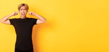 Asyalı rahatsız ve rahatsız edici bir adamın portresi. Surat asıyor, kulaklarını kapatıyor. Gürültüden rahatsız oluyor, sarı arka planda duruyor..