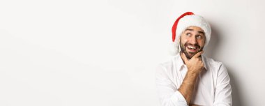 Parti, kış tatili ve kutlama konsepti. Mutlu adamın Noel hediyesi listesi yakın plan, Noel Baba şapkası takıyor, sol üst köşeye bakıyor düşünceli, beyaz arka plan.